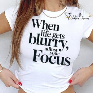 May include: White t-shirt with the text "When life gets blurry, adjust your focus" in black. The shirt has short sleeves and a crew neck. A gold chain necklace and bracelet are also visible.