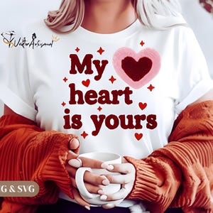 Puede incluir: Camiseta blanca con la frase "My heart is yours" en letras granates, con un corazón rosa con centro rojo oscuro. Corazones rojos y estrellas fugaces acentúan el diseño. La persona sostiene una taza blanca.