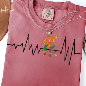 Op de afbeelding: Stofroze Comfort Colors T-shirt met een zwart hartslaglijnontwerp. Een cartoon pretzel-personage rent langs de lijn, met groene en gele accenten. Een zilveren ketting is over het shirt gedrapeerd.