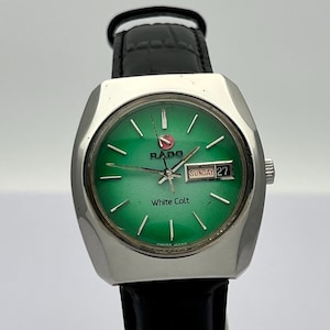 May include: A Rado White Colt wristwatch featuring a silver-tone case, a vibrant green dial with silver hour markers, and a black leather strap. The dial displays the Rado logo, the text White Colt, and a day-date window showing Sunday 27.