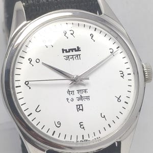 May include: A silver wristwatch with a white face and black fabric band. The watch face features black numerals, text in an Indian language, and silver hands. The watch is a classic design.