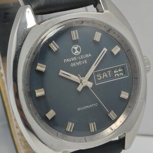 May include: A vintage Favre-Leuba Geneve Duomatic watch with a dark blue face, silver hands, and a day-date display. The watch has a silver case and a black leather strap. The watch face features the brand name and the word "Duomatic".