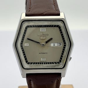 May include: A Seiko 5 automatic wristwatch with a hexagonal silver-tone case and a light grey dial. The watch features black hour and minute hands, a day-date window at the three o'clock position, and a brown leather strap with a textured finish.