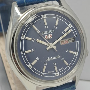 May include: A silver-toned Seiko automatic wristwatch with a navy blue face and a blue leather strap. The watch face displays the day and date, with white numerals and hands. The watch is a classic design.