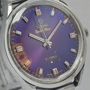 Op de afbeelding: Vintage T.H.O.N.I Space Star polshorloge met een zilverkleurige metalen band en kast. De wijzerplaat is diep paars met zilveren wijzers en markeringen. De wijzerplaat heeft ook de tekst "21 Jewels" en "Swiss Made".