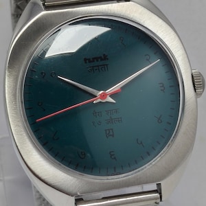 May include: A silver-toned wristwatch with a teal face and a mesh band. The watch face features red hands and Arabic numerals. The watch face has text in a foreign language.