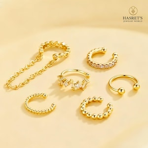 May include: A collection of gold-colored ear cuffs in various designs. One cuff has a chain, others feature beaded, gem-studded, or simple rounded styles. The jewelry is displayed on a light yellow surface, with the brand name "HASRET'S JEWELRY WORLD" visible.