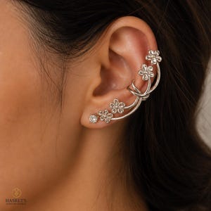 May include: Silver-toned ear cuff featuring a floral design with small, sparkling accents. The cuff curves along the ear, with three flowers clustered together and a single, larger gem at the end. The jewelry is designed to be worn on the ear.
