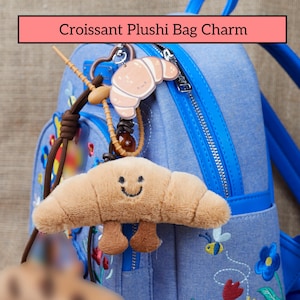 Può includere: Un ciondolo a forma di croissant in peluche marrone chiaro con un volto sorridente e piccole gambe. Il ciondolo è attaccato a uno zaino blu. Il testo "Croissant Plushi Bag Charm" è in un rettangolo rosa in alto.