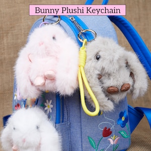 Könnte beinhalten: Eine Sammlung von Plüsch-Hasen-Schlüsselanhängern in Rosa, Grau und Weiß, die an einem blauen Jeans-Rucksack befestigt sind. Die Schlüsselanhänger haben flauschiges Fell und sind mit gelben und lila Riemen versehen. Der Text "Bunny Plushi Keychain" wird oben angezeigt.