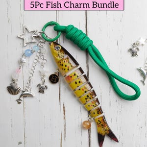 May include: A fish-shaped charm bundle with a green cord and various charms. The fish charm is yellow and brown with a segmented design. Additional charms include a star, seashell, dolphin, and turtle. The text "5Pc Fish Charm Bundle" is in a pink box.