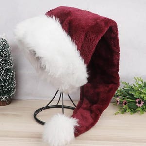 Könnte beinhalten: Eine lange, rot-weiße Weihnachtsmannmütze mit einem flauschigen weißen Bommel am Ende. Die Mütze ist aus weichem, plüschem Stoff gefertigt und perfekt, um jeder Weihnachtsdekoration eine festliche Note zu verleihen.