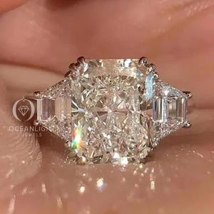 Puede incluir: Un radiante anillo de compromiso de platino con un gran diamante central de talla rectangular y dos diamantes laterales de talla esmeralda. El anillo brilla con facetas brillantes. El logotipo de Oceanlight Jewels es visible.