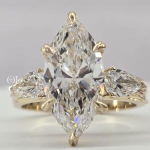 Puede incluir: Un anillo de compromiso con diamantes talla marquesa y una banda de oro. El anillo presenta un gran diamante central talla marquesa y dos diamantes más pequeños en forma de pera. La banda tiene detalles intrincados.