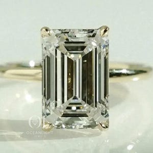May include: An emerald-cut diamond ring with a gold band. The diamond is rectangular with step-cut facets, reflecting light. The ring is set on a white surface, with the Oceanlight Jewels logo visible.