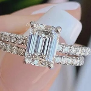 May include: A silver-toned ring featuring a large emerald-cut center stone held by four prongs, flanked by a band encrusted with small round pav&eacute;-set stones. The metal band displays a bright, reflective finish with clear, faceted gemstones.
