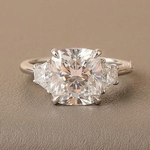 Puede incluir: Un anillo de compromiso plateado con una piedra central grande de corte cuadrado y dos piedras laterales triangulares más pequeñas. El anillo está sobre una superficie de tono neutro, con un fondo suave y borroso.