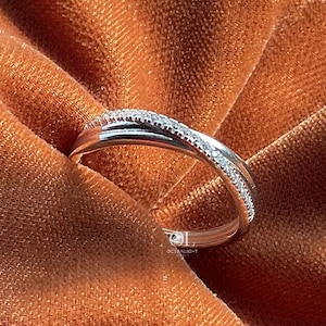 Puede incluir: Un anillo plateado con un dise&ntilde;o entrecruzado, engastado con peque&ntilde;as piedras preciosas transparentes. El anillo se exhibe sobre una tela texturizada de color &oacute;xido, creando un contraste entre el metal brillante y el fondo mate. El dise&ntilde;o del anillo es elegante y moderno.