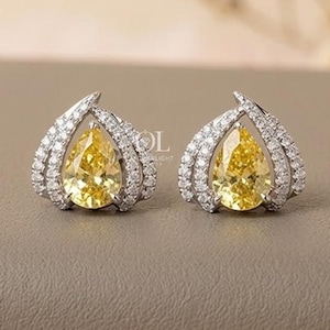 May include: A pair of stud earrings featuring a central pear-cut yellow gemstone surrounded by a silver-toned frame encrusted with small clear white stones. The design has a pointed top and a curved shape, with the text OL visible in the center.