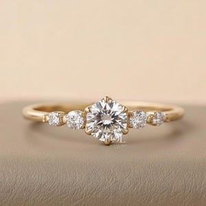 May include: A gold ring with a large, round, clear gemstone in the center, surrounded by smaller, clear gemstones. The ring is set on a neutral surface with a soft, pink background.