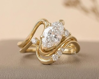 May include: A gold ring featuring a central marquise-cut clear gemstone flanked by smaller round-cut stones. The band has a fluid, asymmetrical design that curves around the center setting, creating a layered appearance with polished gold metal.