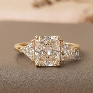 Puede incluir: Un anillo de oro con un gran diamante central de talla rectangular, flanqueado por dos diamantes más pequeños de talla triangular. El anillo tiene un diseño clásico y elegante, ideal para compromisos u ocasiones especiales.