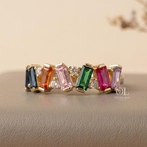 Può includere: Un anello d'oro con una fila di pietre rettangolari colorate nei toni del blu, arancione, rosa, verde e viola. Piccole pietre bianche sono intervallate tra le pietre più grandi. L'anello è appoggiato su una superficie neutra.