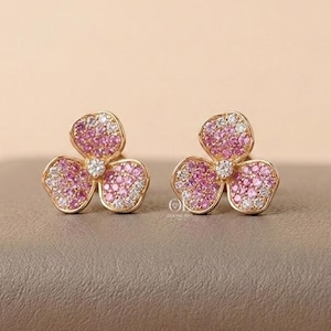 May include: A pair of gold stud earrings shaped like three-petaled flowers. Each petal is encrusted with small pink and white stones, with a single clear stone at the center of each flower. The earrings feature a polished gold metal finish.