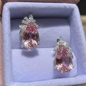 May include: A pair of elegant drop earrings displayed in a pink jewelry box. Each earring features a large, pear-shaped pink gemstone, accented with smaller, sparkling white stones. The earrings are set in a silver-colored metal.