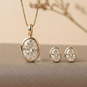 May include: A gold necklace featuring an oval-shaped clear gemstone pendant in a bezel setting, accompanied by a pair of matching oval-shaped stud earrings. The jewelry set displays a polished gold finish and clear faceted stones throughout.
