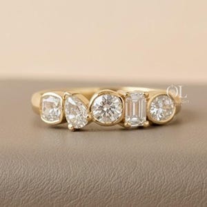 May include: A gold ring featuring five clear gemstones of varying shapes: rectangular, pear, round, emerald cut, and round. The ring is set on a neutral-toned surface, with a soft, pinkish-white background.