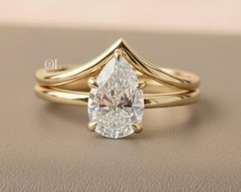 May include: A gold engagement ring set featuring a pear-shaped diamond center stone. The ring is paired with a chevron-shaped band. The set is displayed on a neutral surface with a soft, blurred background.