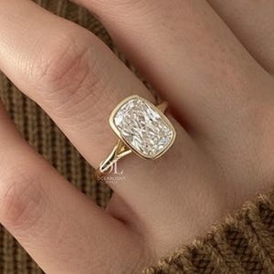 May include: A gold ring with a large, rectangular, clear gemstone. The ring has a simple, elegant design with a thin band and a bezel setting. The gemstone is set in a way that maximizes its brilliance.