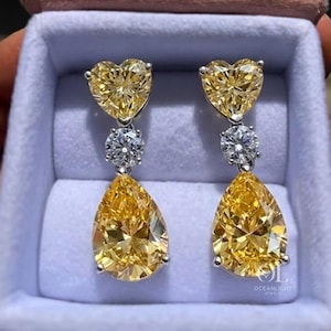May include: A pair of elegant drop earrings featuring heart-shaped and pear-shaped yellow gemstones. The earrings are set in a silver metal and presented in a light blue jewelry box with a pink velvet lid. The earrings are a luxurious accessory.