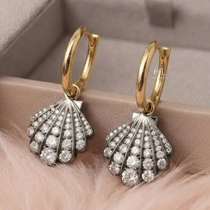May include: A pair of gold hoop earrings featuring dangling seashell-shaped charms. Each shell charm is crafted from silver-toned metal and is encrusted with numerous small, clear, round-cut stones arranged in a radiating fan pattern.