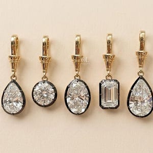 May include: Five gold pendants featuring clear faceted gemstones in various shapes including pear, round, oval, and emerald cuts. Each stone is set in a dark metal frame and suspended from a gold bail. The text OL is visible on the center pendant.