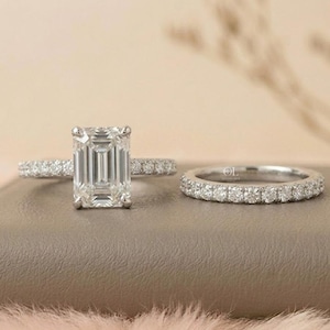 May include: A two-piece ring set featuring a large emerald-cut diamond engagement ring and a matching diamond band. The rings are set in a silver metal, with small diamonds lining the band of the engagement ring and the entire band of the wedding ring.