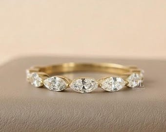 May include: A gold band ring featuring a series of marquise-cut gemstones. The ring is set on a neutral surface with a soft, blurred background. The ring's design is elegant and eye-catching, suitable for various occasions.