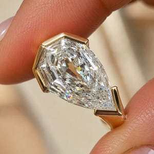 Puede incluir: Un anillo de oro con una gran piedra preciosa transparente en forma de pera, tallada con facetas y montada en un engaste parcial. La banda de oro amarillo pulido muestra múltiples facetas triangulares que reflejan la luz en su superficie.