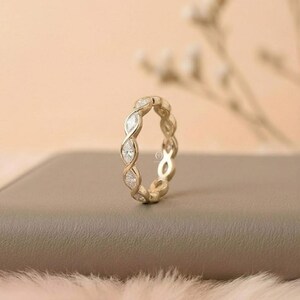 May include: A gold-colored eternity ring with alternating marquise-cut diamonds. The ring is set on a neutral-toned surface, with a soft, blurred background. The design features a continuous pattern of diamonds, creating a sparkling effect.
