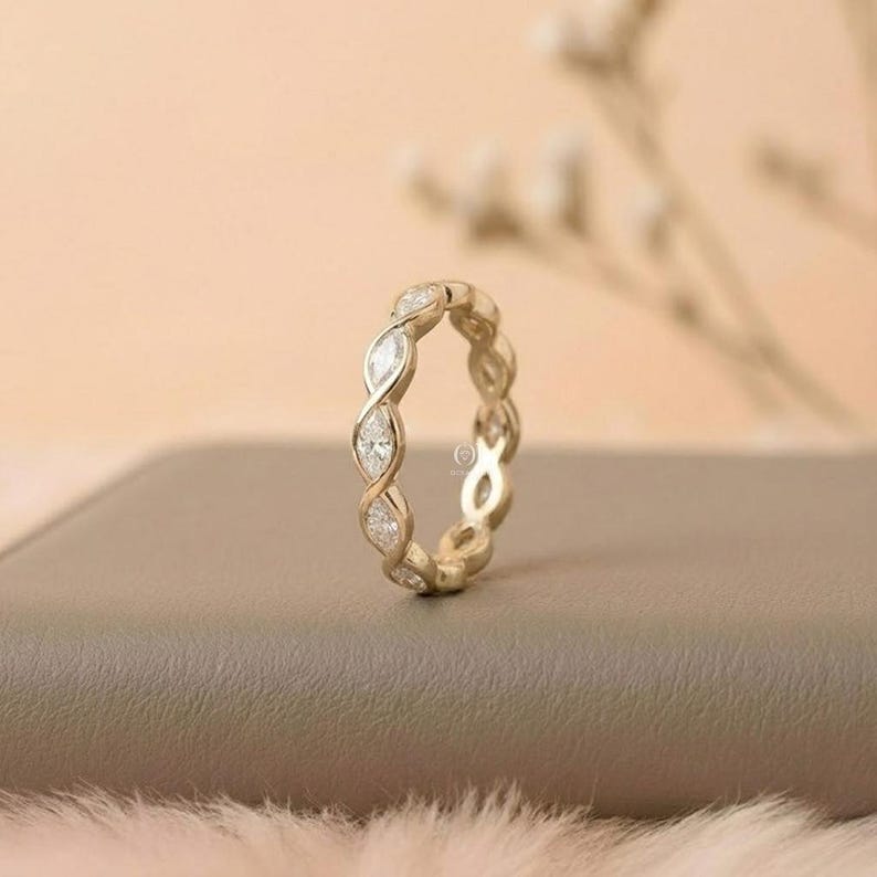 May include: A gold-colored eternity ring with alternating marquise-cut diamonds. The ring is set on a neutral-toned surface, with a soft, blurred background. The design features a continuous pattern of diamonds, creating a sparkling effect.