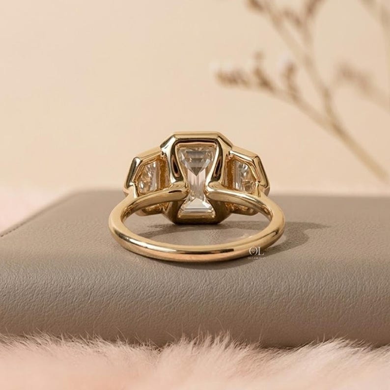 Puede incluir: Un anillo de oro con una gran piedra central de corte esmeralda flanqueada por dos piedras laterales m&aacute;s peque&ntilde;as de corte trapezoide. La banda es de oro pulido y el engaste muestra un dise&ntilde;o claro y multifac&eacute;tico con garras.