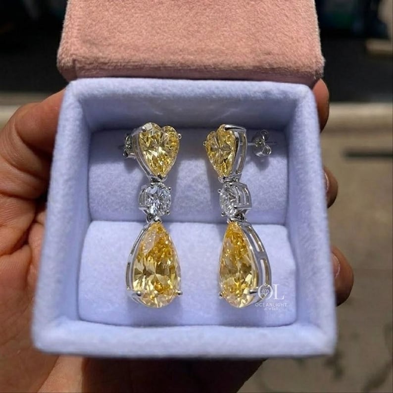 May include: A pair of elegant drop earrings featuring heart-shaped and pear-shaped yellow gemstones. The earrings are set in a silver metal and presented in a light blue jewelry box with a pink lid.