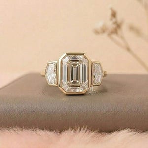Puede incluir: Un anillo de oro con una gran piedra central de corte esmeralda flanqueada por dos piedras laterales m&aacute;s peque&ntilde;as de corte escudo. La banda de oro amarillo pulido sostiene las tres piedras transparentes y facetadas en un engaste de bisel.