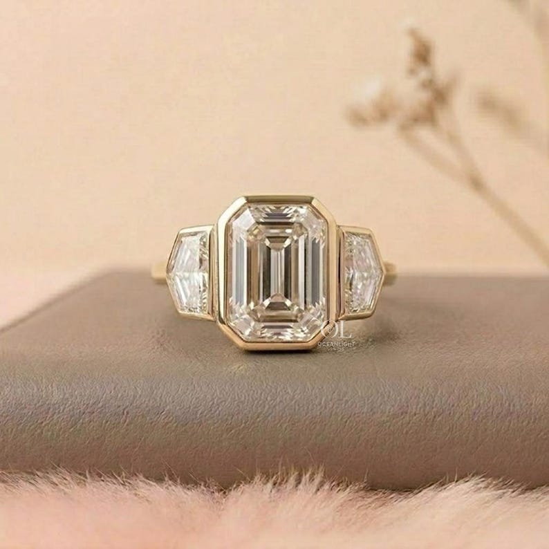 Puede incluir: Un anillo de oro con una gran piedra central de corte esmeralda flanqueada por dos piedras laterales m&aacute;s peque&ntilde;as de corte escudo. La banda de oro amarillo pulido sostiene las tres piedras transparentes y facetadas en un engaste de bisel.