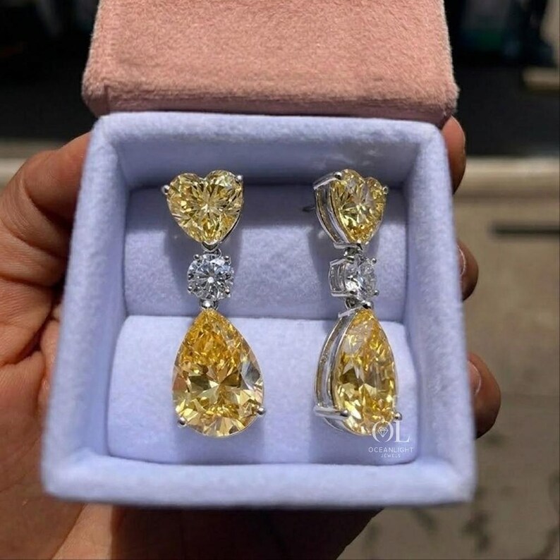 May include: A pair of elegant earrings with a heart-shaped yellow gemstone at the top, a round white gemstone in the middle, and a pear-shaped yellow gemstone at the bottom. The earrings are displayed in a light blue jewelry box with a pink lid.