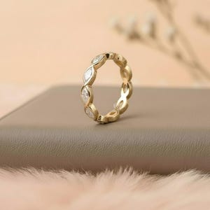 May include: A gold ring with a repeating pattern of marquise-cut gemstones. The ring is set with clear, sparkling stones and has a polished, yellow gold band. The ring is displayed on a neutral-toned surface.