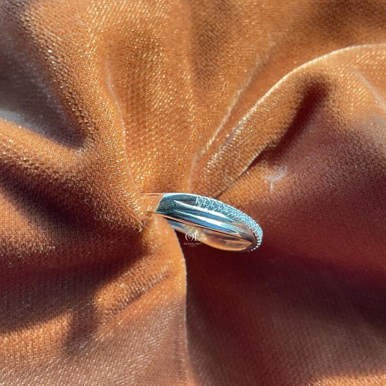 Puede incluir: Un anillo plateado con una parte superior clara y rectangular y una banda incrustada con peque&ntilde;as piedras brillantes. El anillo se coloca sobre un fondo de tela suave, texturizada, de color marr&oacute;n anaranjado.