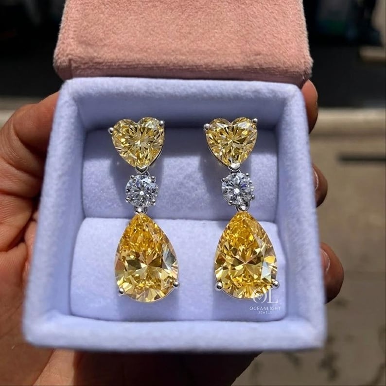 May include: A pair of elegant drop earrings featuring heart-shaped and pear-shaped yellow gemstones. The earrings are set in a silver metal and presented in a light blue jewelry box with a pink velvet lid. The earrings are a luxurious accessory.
