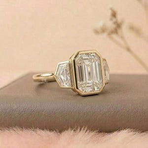 Puede incluir: Un anillo de oro con una gran piedra preciosa rectangular y transparente flanqueada por dos piedras preciosas m&aacute;s peque&ntilde;as de forma geom&eacute;trica. El anillo est&aacute; sobre una superficie de tono neutro, con un fondo suave y borroso.